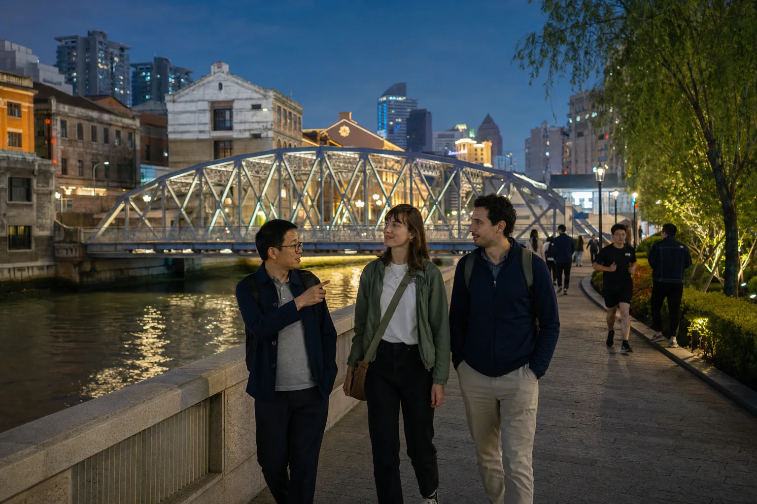 Travelers walking with a local guide beside Suzhou Creek and an illuminated historic bridge in Shanghai